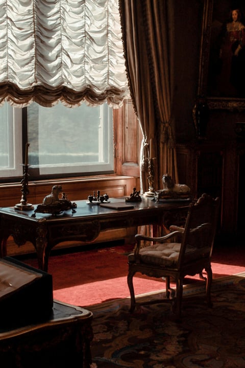 Ornate wooden desk by a window with gathered cream curtains, dark wood chair, and classical interior decor