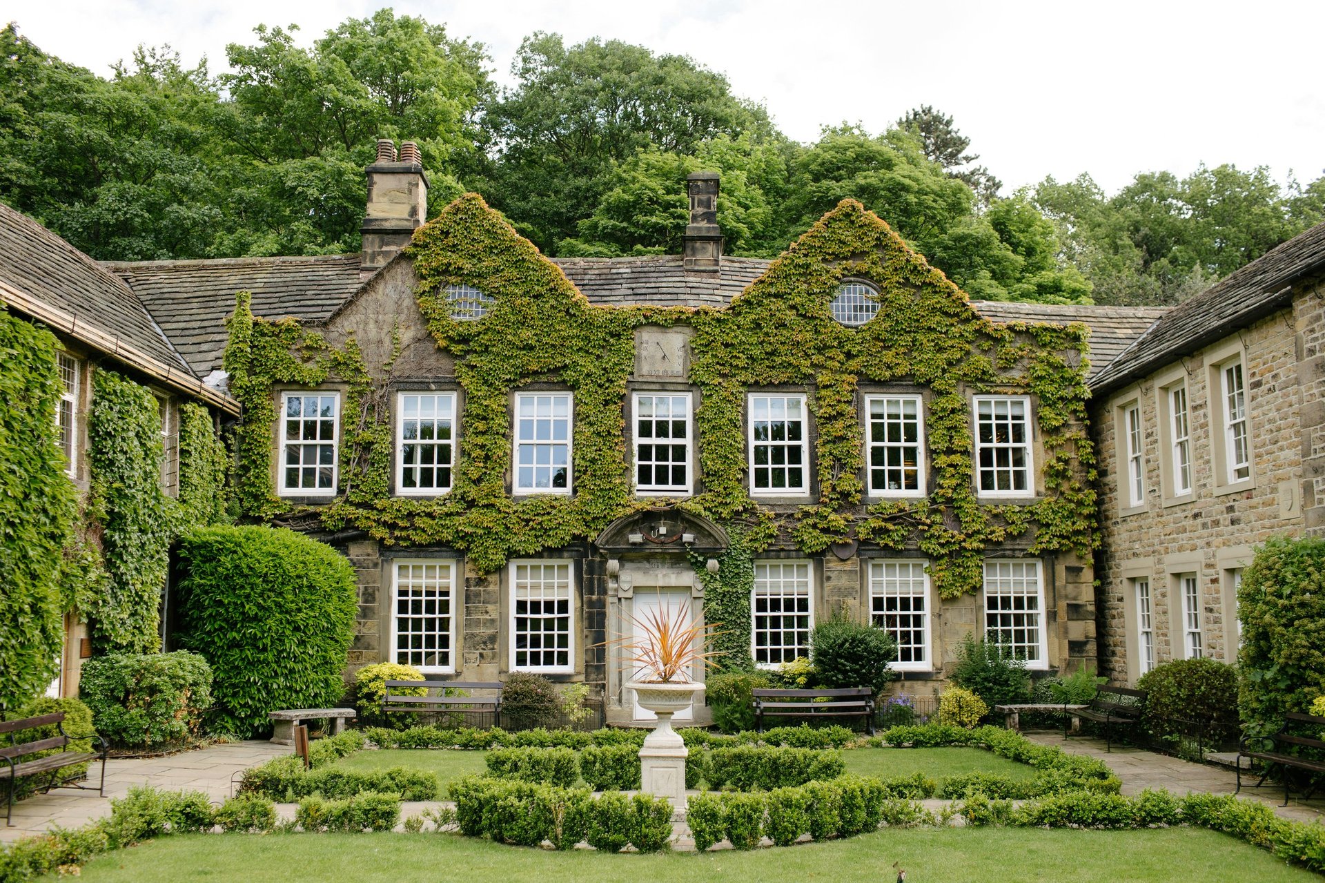 Historic stone mansion covered in ivy with symmetrical windows, formal garden, and manicured hedges in foreground
